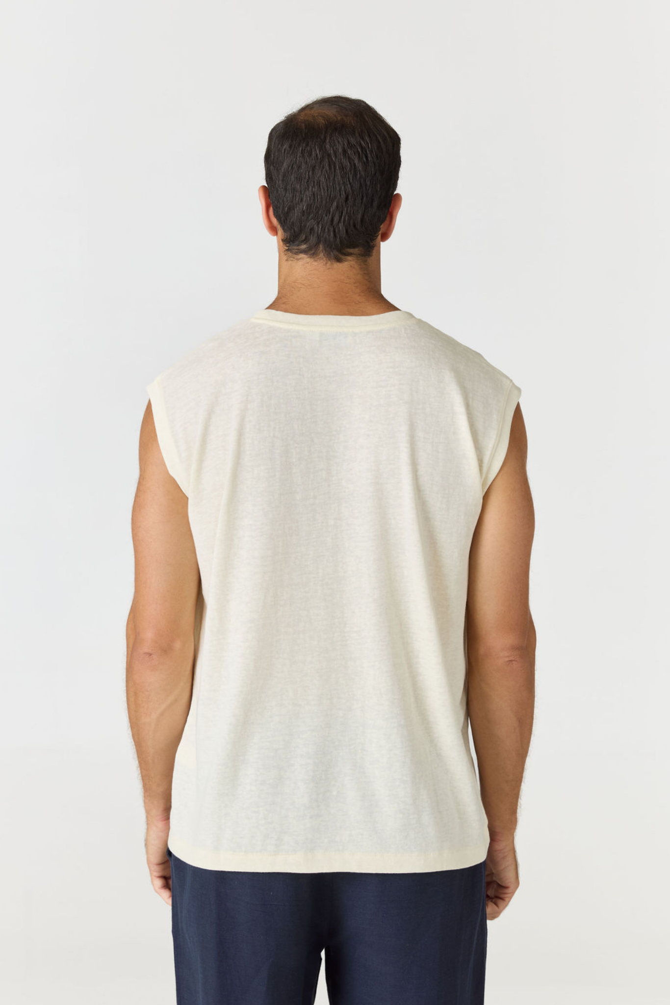 A man viewed from behind wearing a sleeveless cream-colored t-shirt and dark navy pants, standing against a plain light gray studio background.