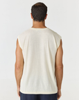 A man viewed from behind wearing a sleeveless cream-colored t-shirt and dark navy pants, standing against a plain light gray studio background.