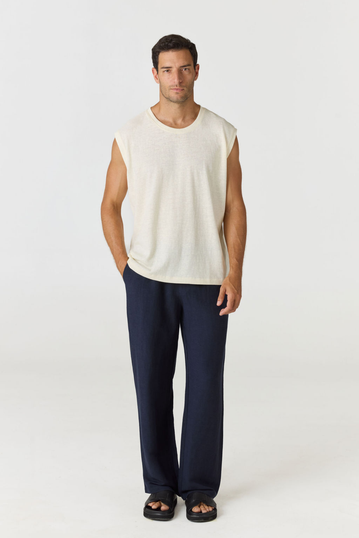 A man modeling a sleeveless cream knit top and navy blue pants against a plain light gray studio background.