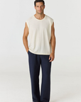 A man modeling a sleeveless cream knit top and navy blue pants against a plain light gray studio background.