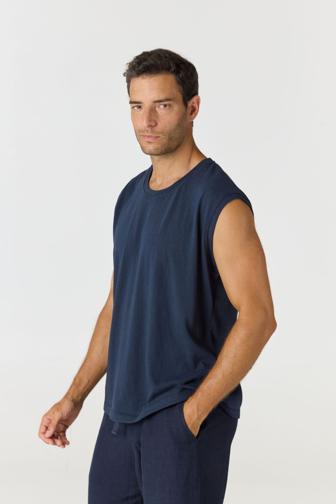 A man wearing a sleeveless navy blue muscle tank top and matching navy pants, standing against a light gray studio background.