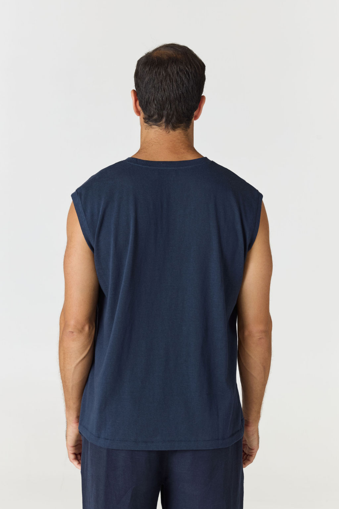 A man viewed from the back wearing a sleeveless navy blue shirt and matching navy pants against a light gray studio background.