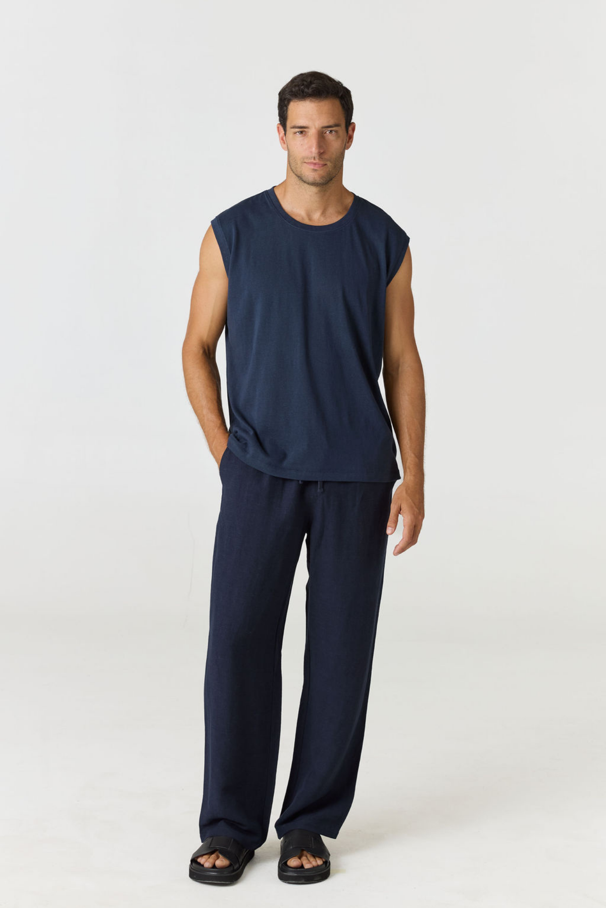 A man modeling a dark navy blue sleeveless top and matching navy blue pants against a plain light gray studio background.