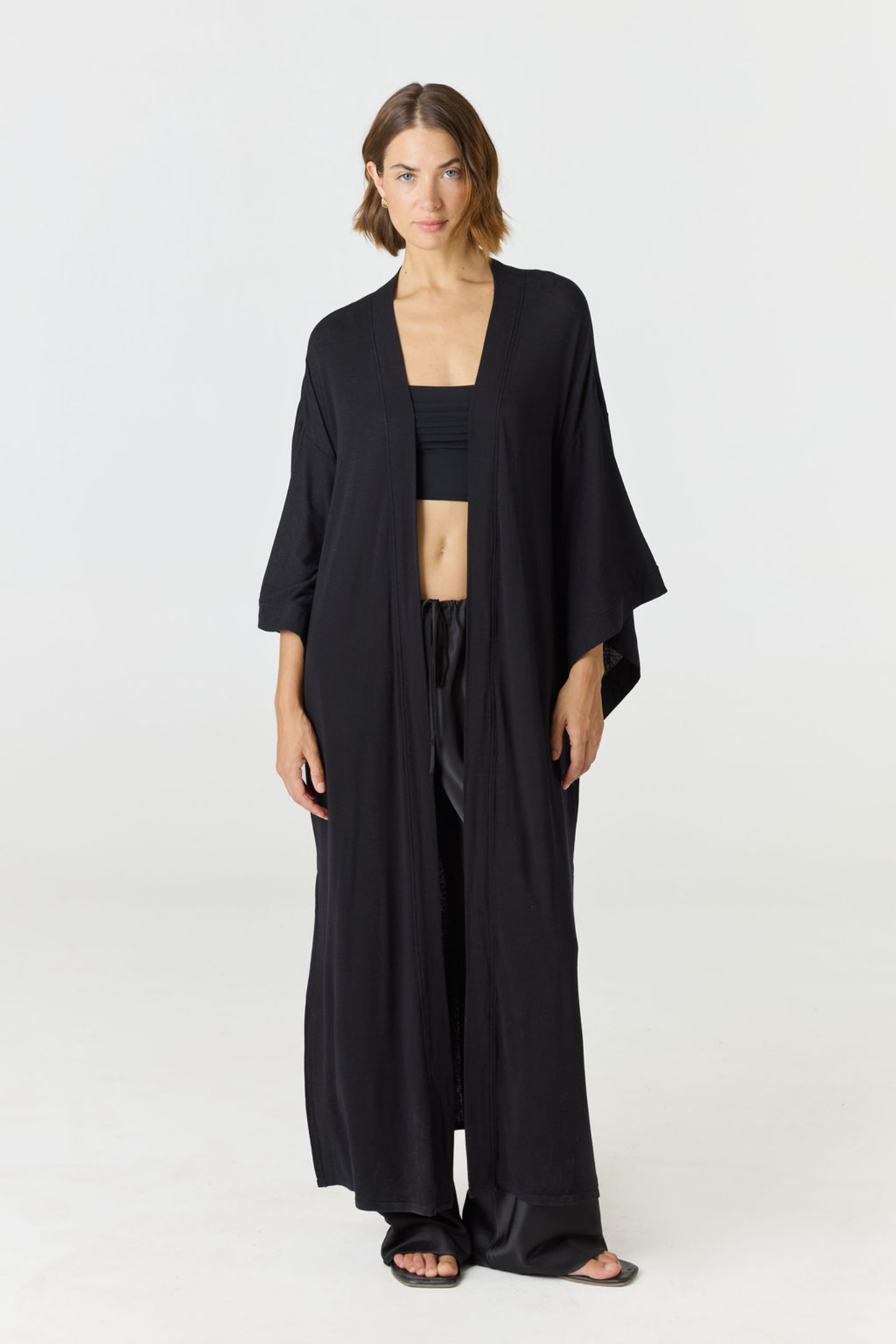 A woman modeling a black open-front kimono-style cardigan over a black crop top and wide-leg pants, photographed against a clean light gray studio background.