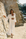 A woman stands on a sandy coastal path between large rock formations, wearing a flowing white long-sleeve cardigan over a matching white crop top and high-waisted skirt. The background shows sunlit stone cliffs, a bit of water, and distant buildings.