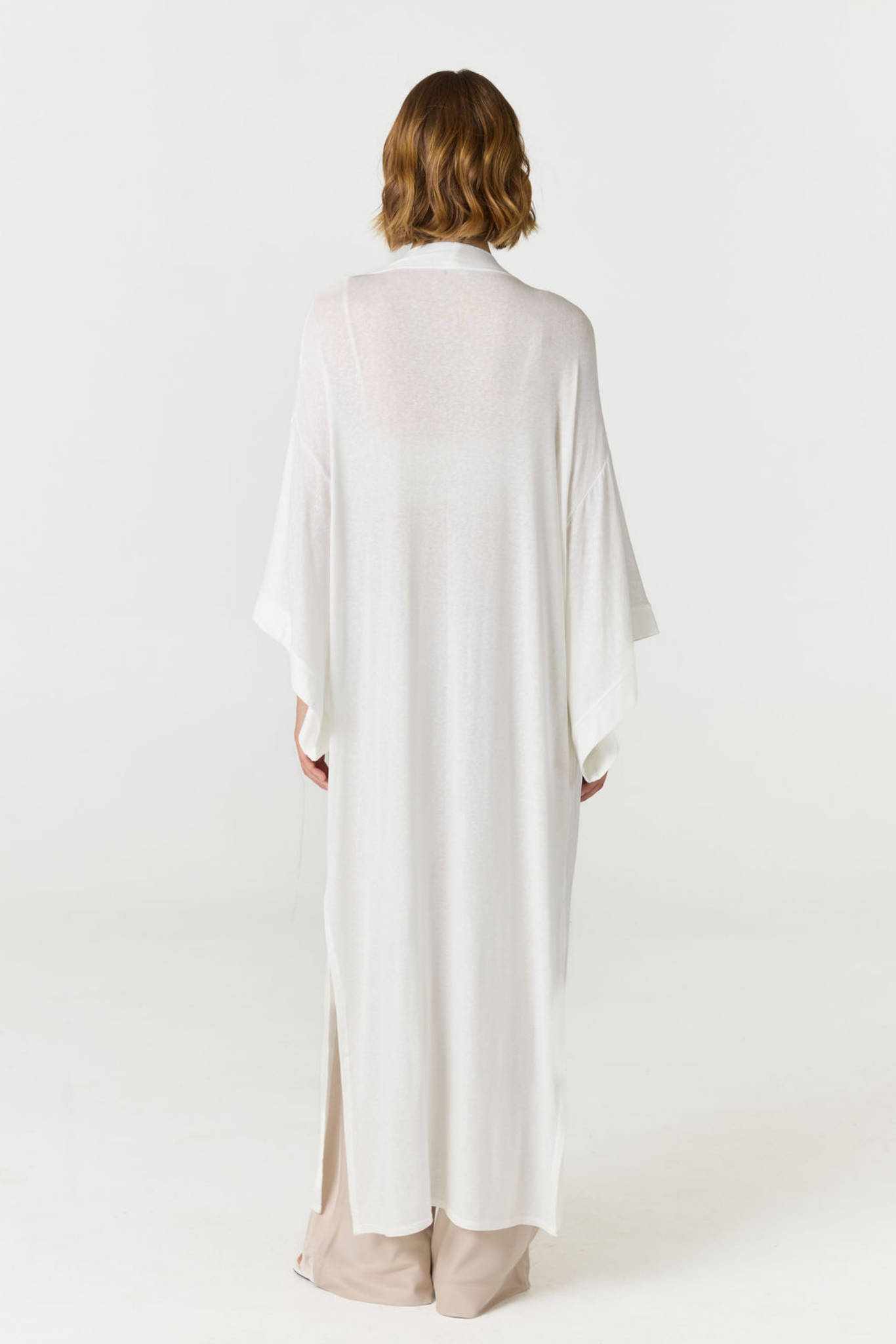 A woman viewed from behind wearing a long, loose white kimono-style top over beige pants, standing against a plain white studio background.
