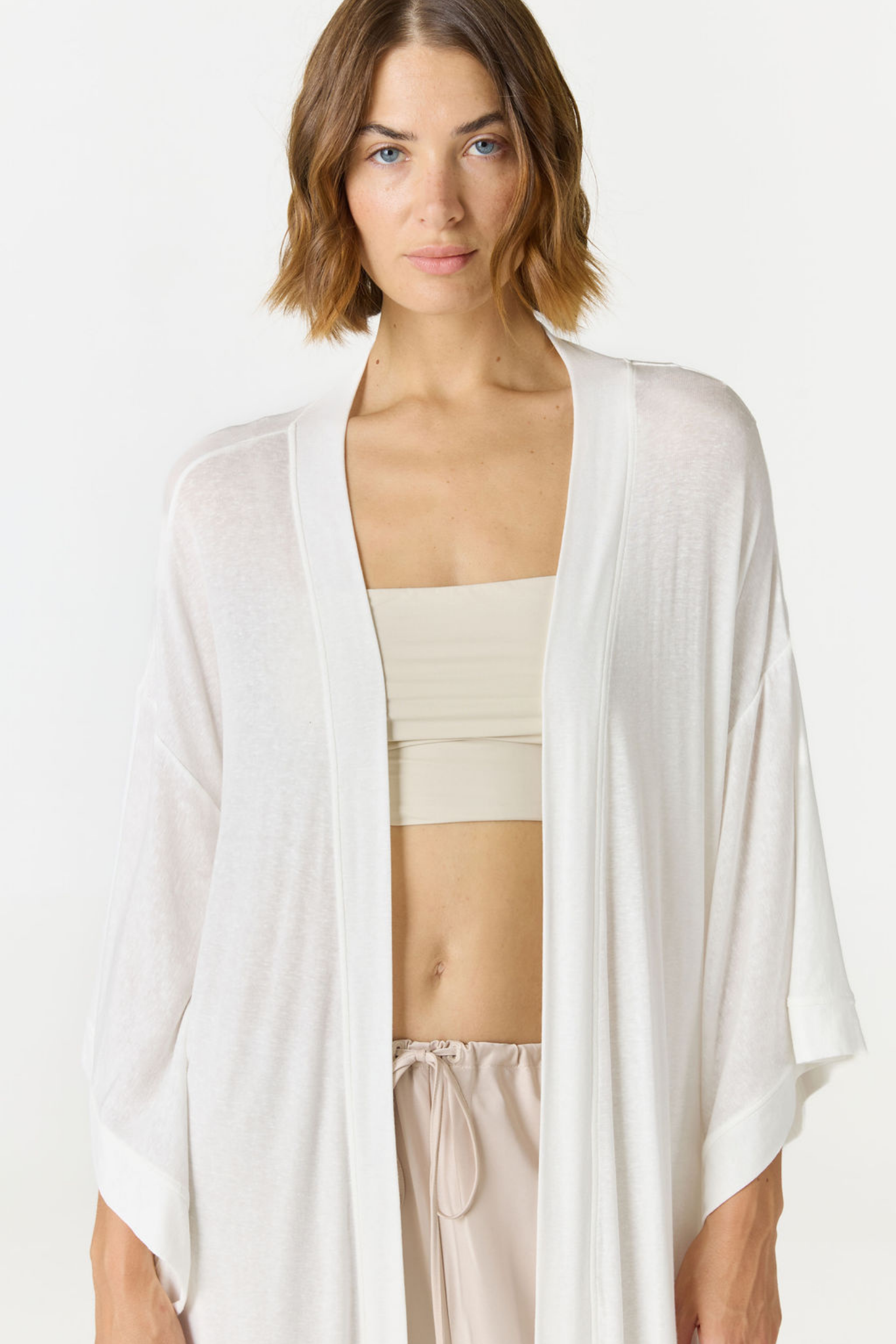 A woman modeling a lightweight, open-front white cardigan over a beige crop top and light drawstring pants against a plain white studio background.