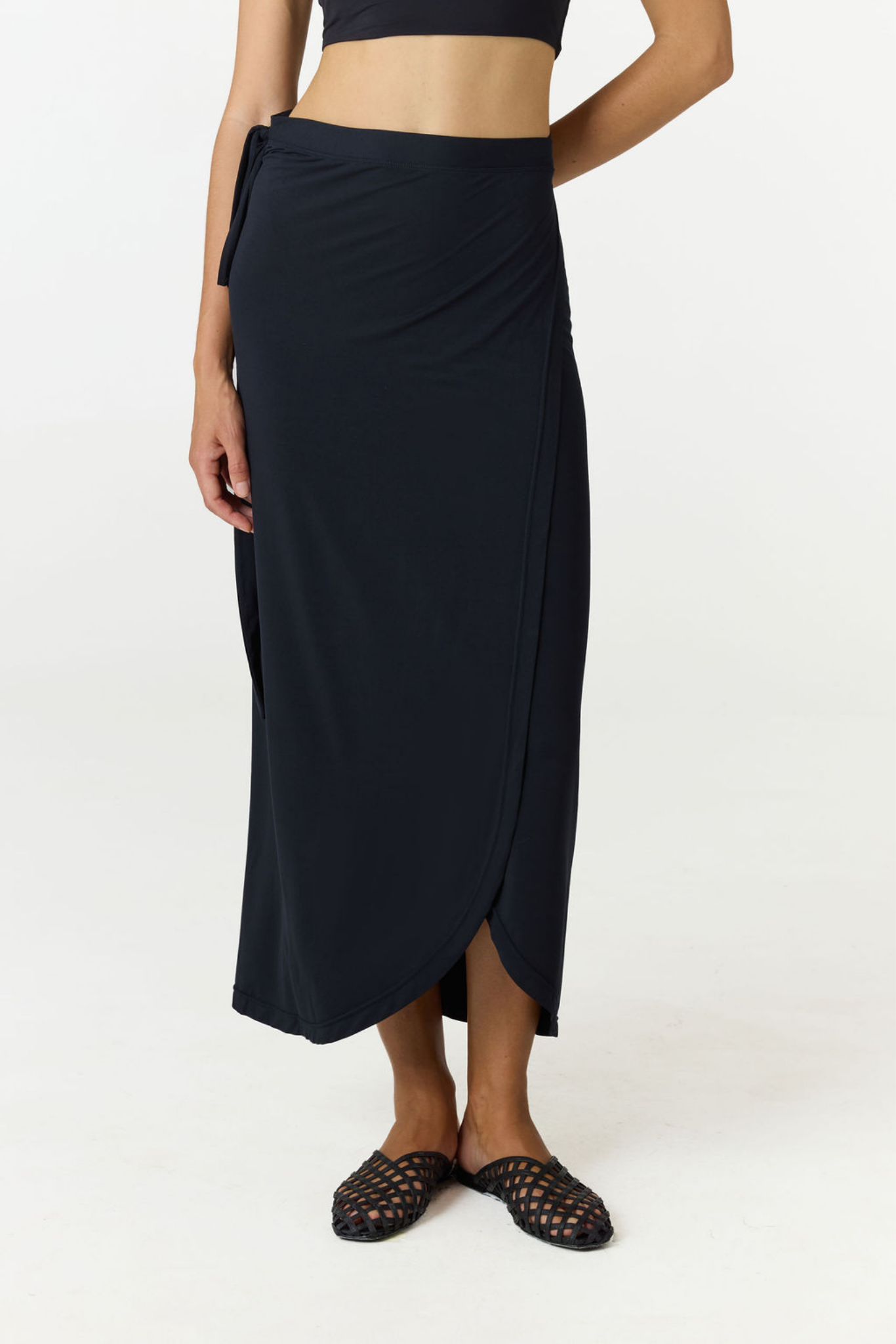 A model wearing a dark navy wrap midi skirt with a side tie and a front slit, paired with black woven slide sandals against a plain white studio background.