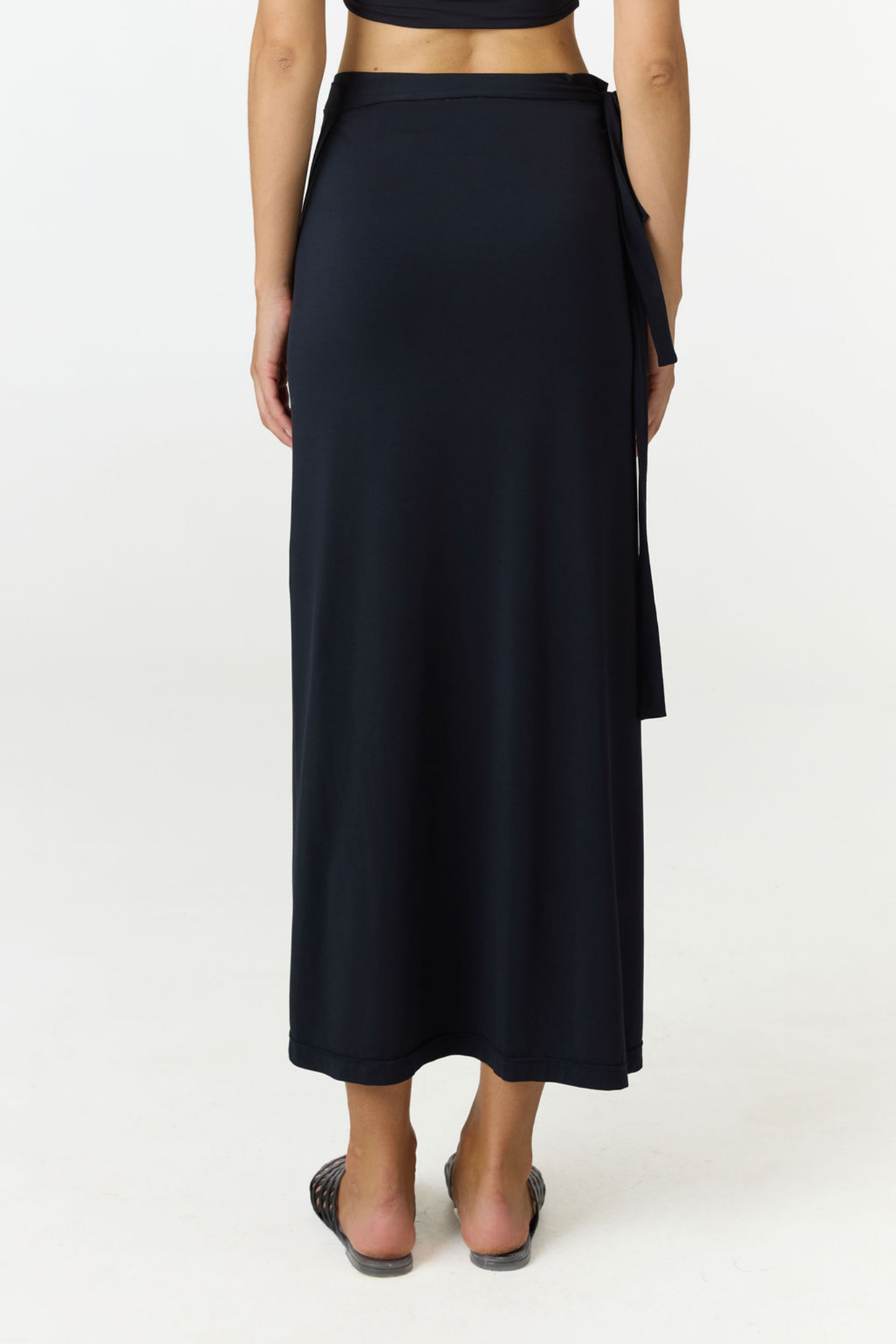 A model shown from the back wearing a dark navy wrap midi skirt with side ties, paired with flat sandals, against a plain white studio background.