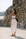 A woman stands outdoors against a rustic stone wall wearing a white two-piece outfit consisting of a sleeveless crop top and a long skirt, with a black shoulder bag and flat sandals.