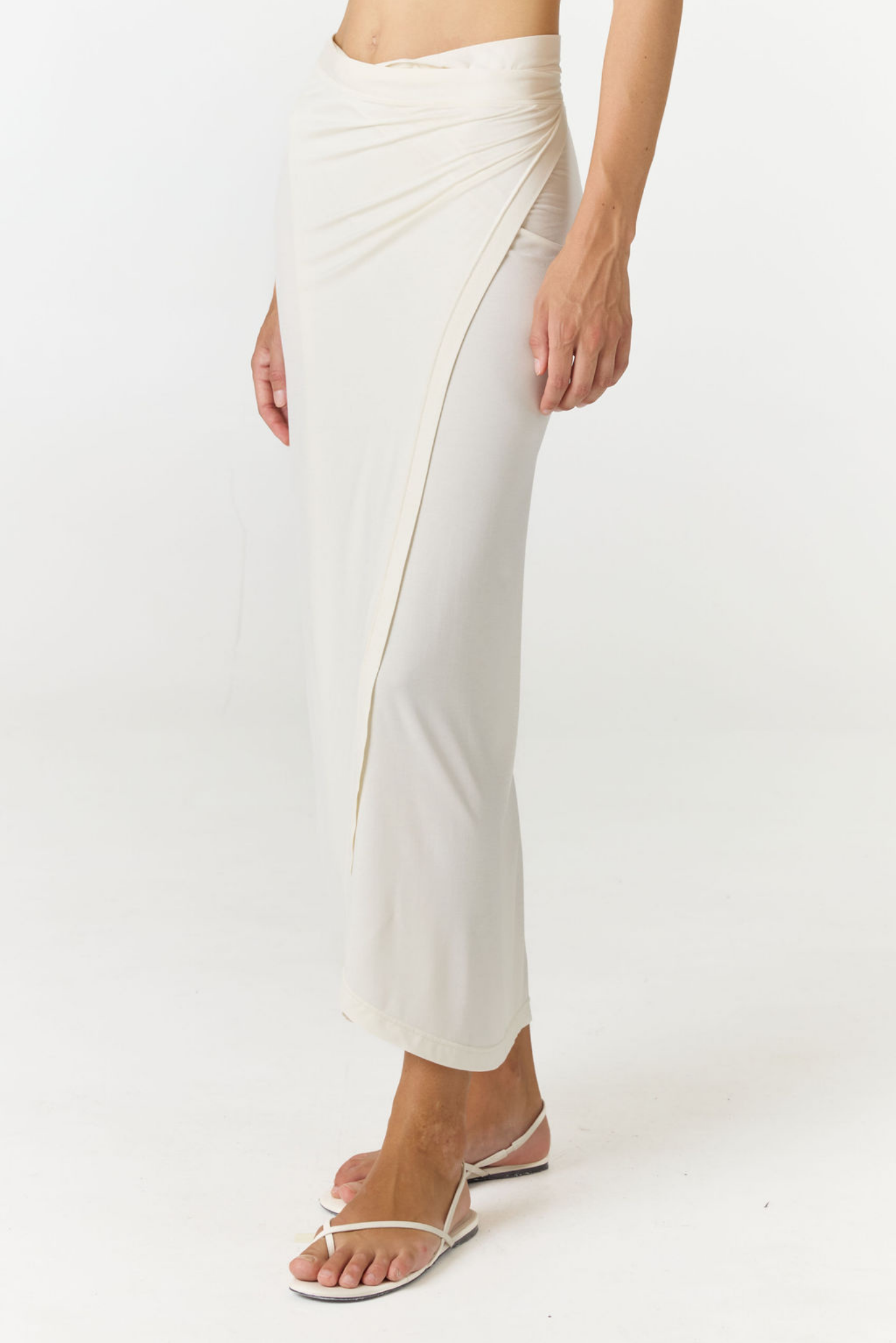 A woman wearing a draped wrap maxi skirt in off-white, paired with white flat sandals, shown from the waist down against a plain white studio background.