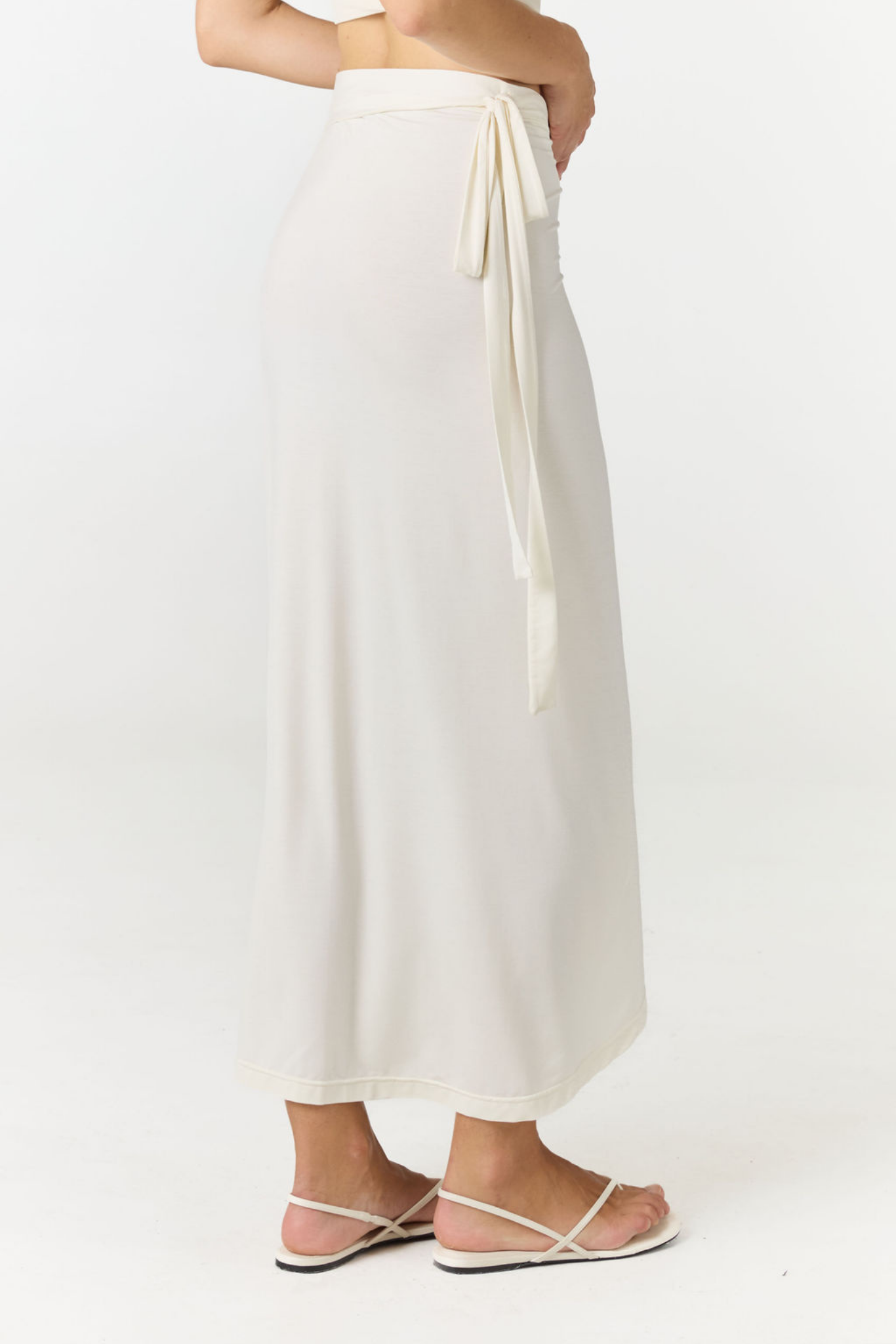 A woman wearing a long off-white maxi skirt with a side tie detail, paired with white strappy flat sandals, shown from the waist down against a plain white studio background.