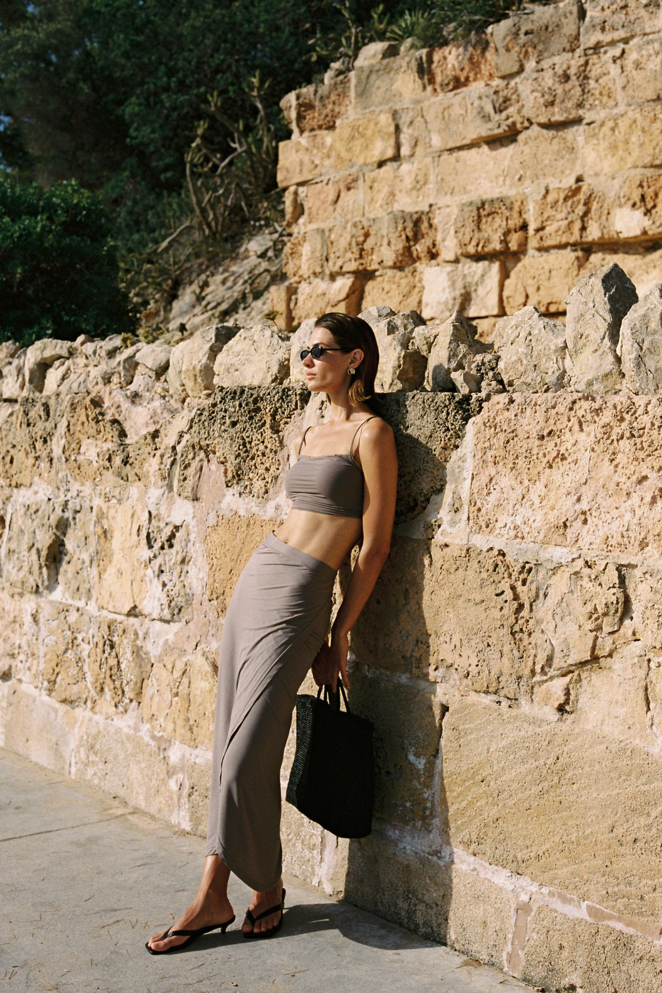 A woman wearing a taupe two-piece set consisting of a cropped tank top and high-waisted wrap skirt, posing against a rustic stone wall.