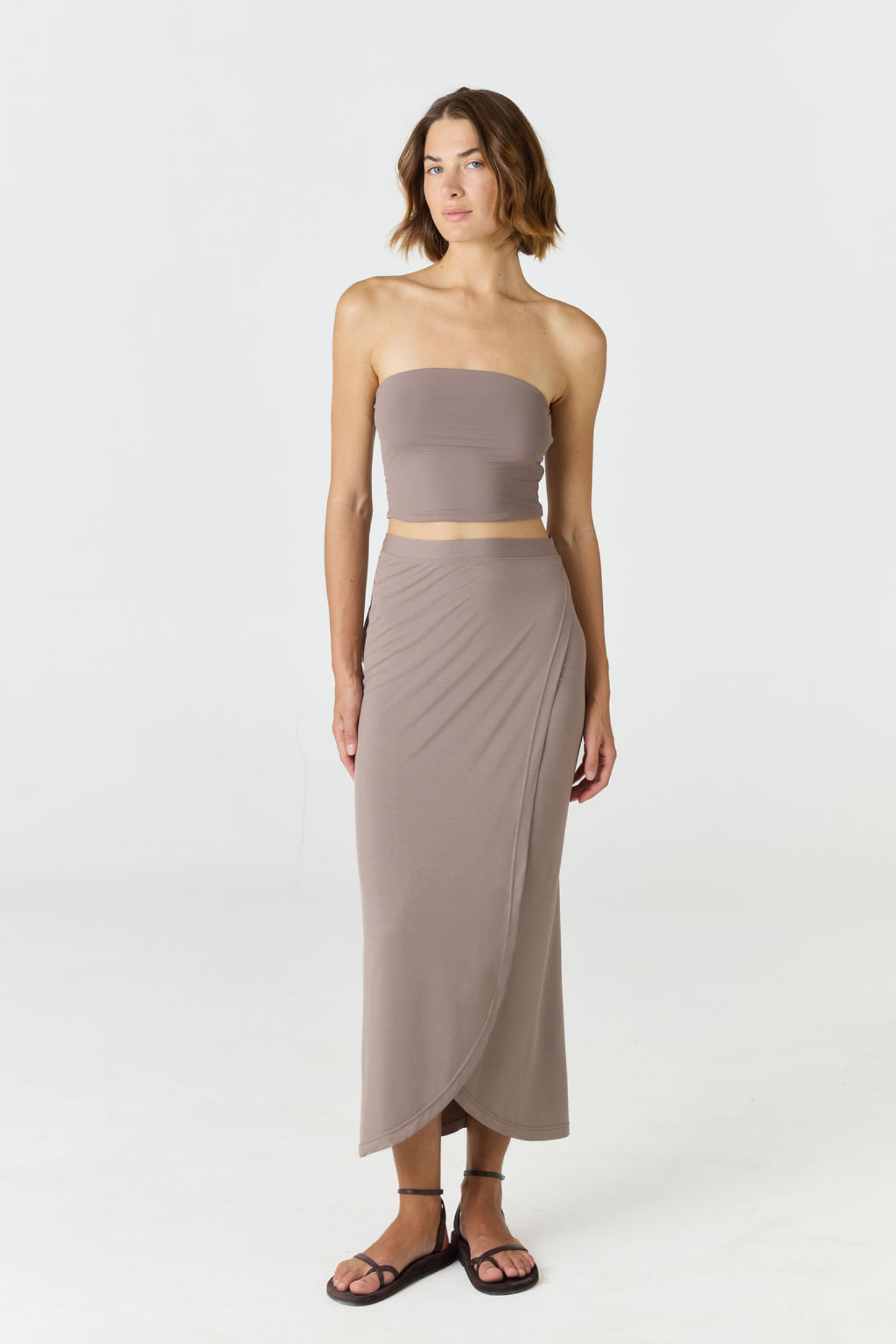 A woman modeling a two-piece matching set consisting of a strapless bandeau crop top and a high-waisted draped midi skirt in a muted taupe-brown color, paired with black strappy sandals against a plain white studio background.