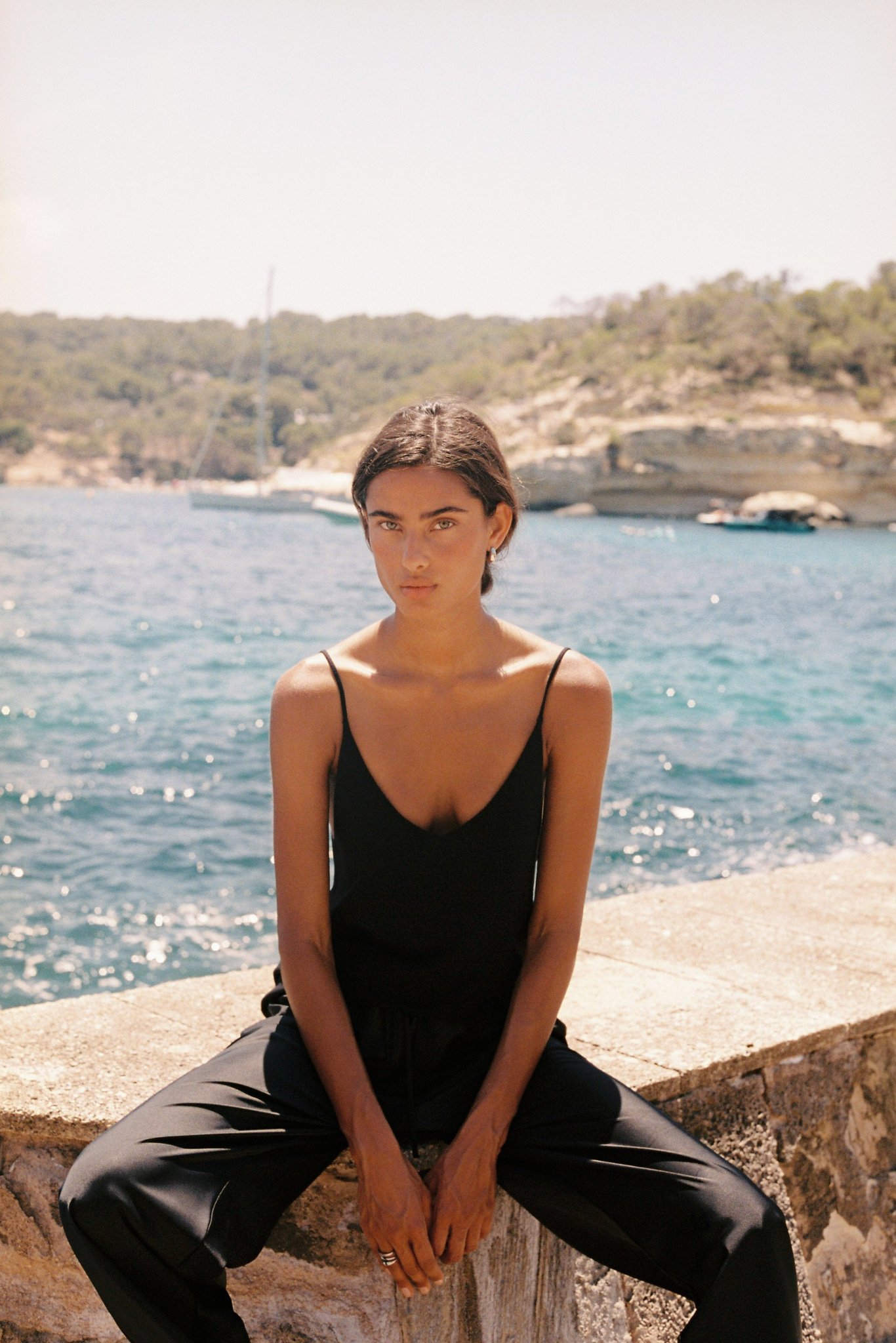 A woman wearing a black spaghetti-strap camisole top and black trousers sits on a stone wall with a sunny blue sea and rocky coastline in the background.