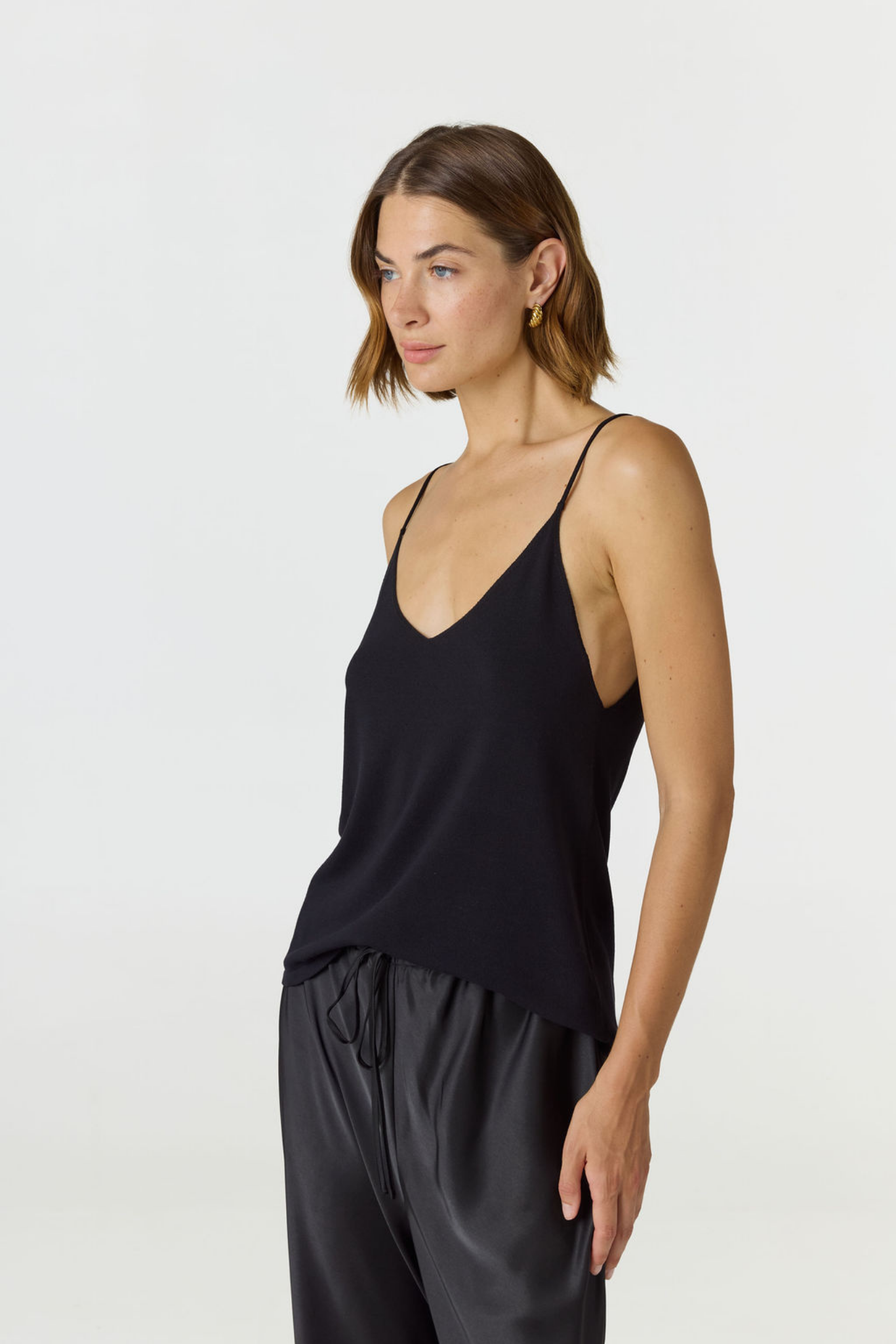 A woman wearing a black spaghetti-strap camisole top and black satin pants, photographed against a clean light gray studio background.