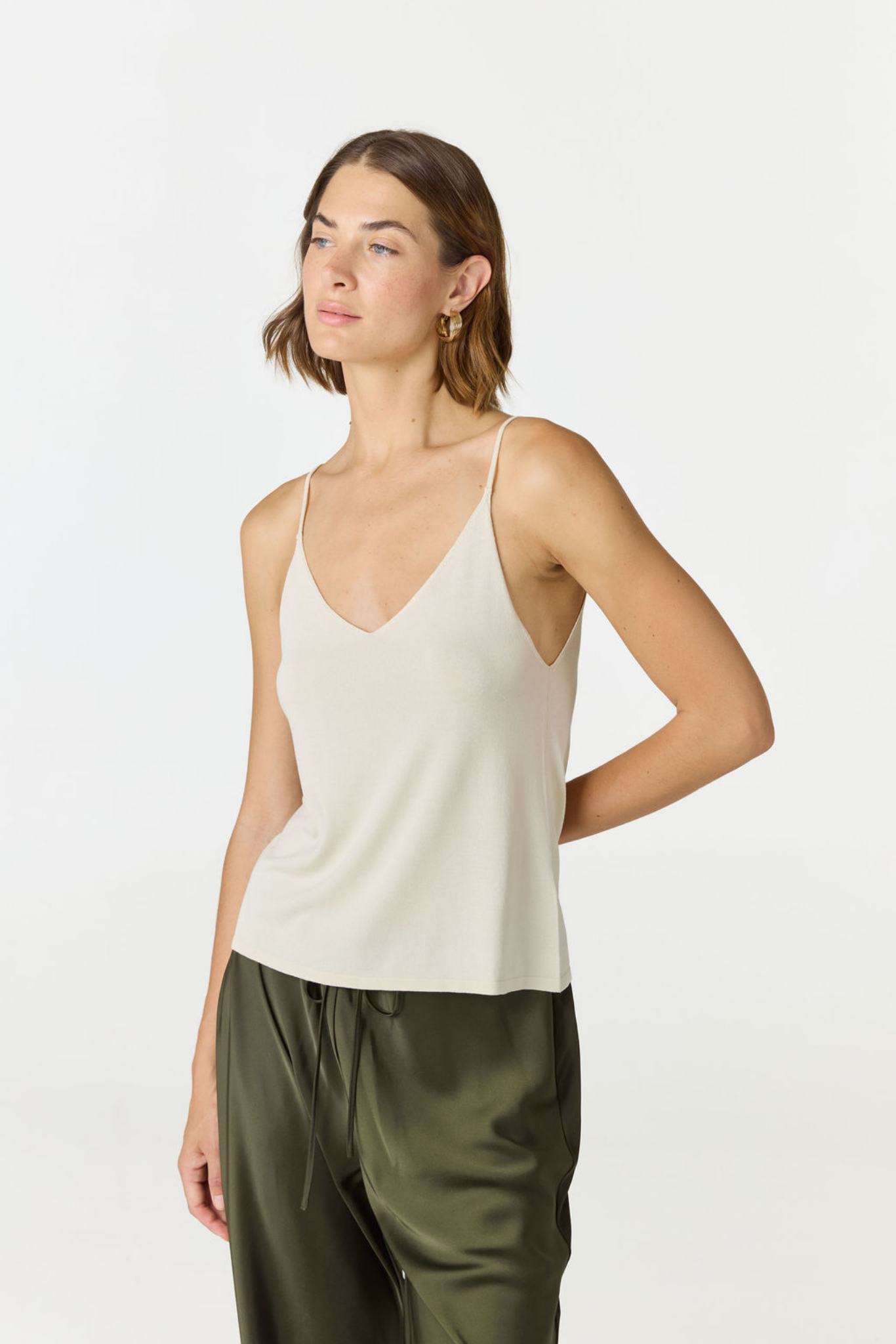 A woman modeling a light beige spaghetti-strap camisole top paired with olive green satin pants, photographed against a clean white studio background.